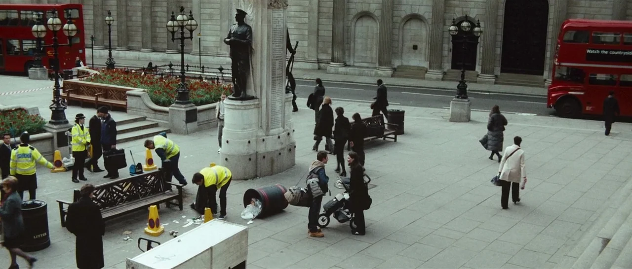 Green Street (2005) movie still: wide — A stone city plaza features a central war memorial monument flanked by ornate lanterns. Pedestrians navigate…