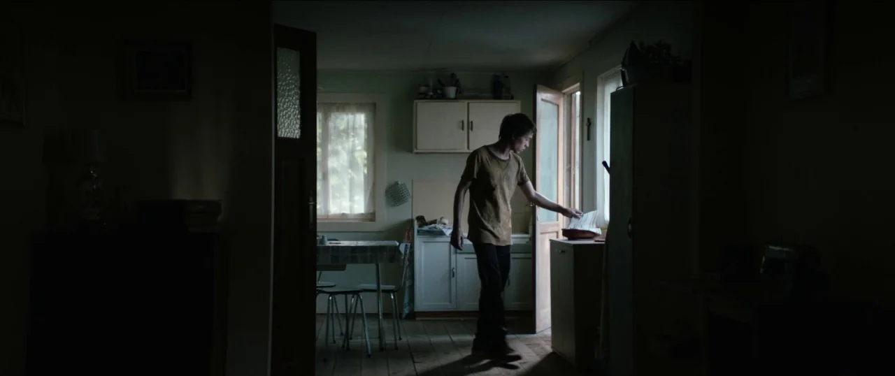 Good Favour (2017) movie still: medium — A dimly lit room features wooden flooring and light-colored cabinetry against the back wall. A young man…
