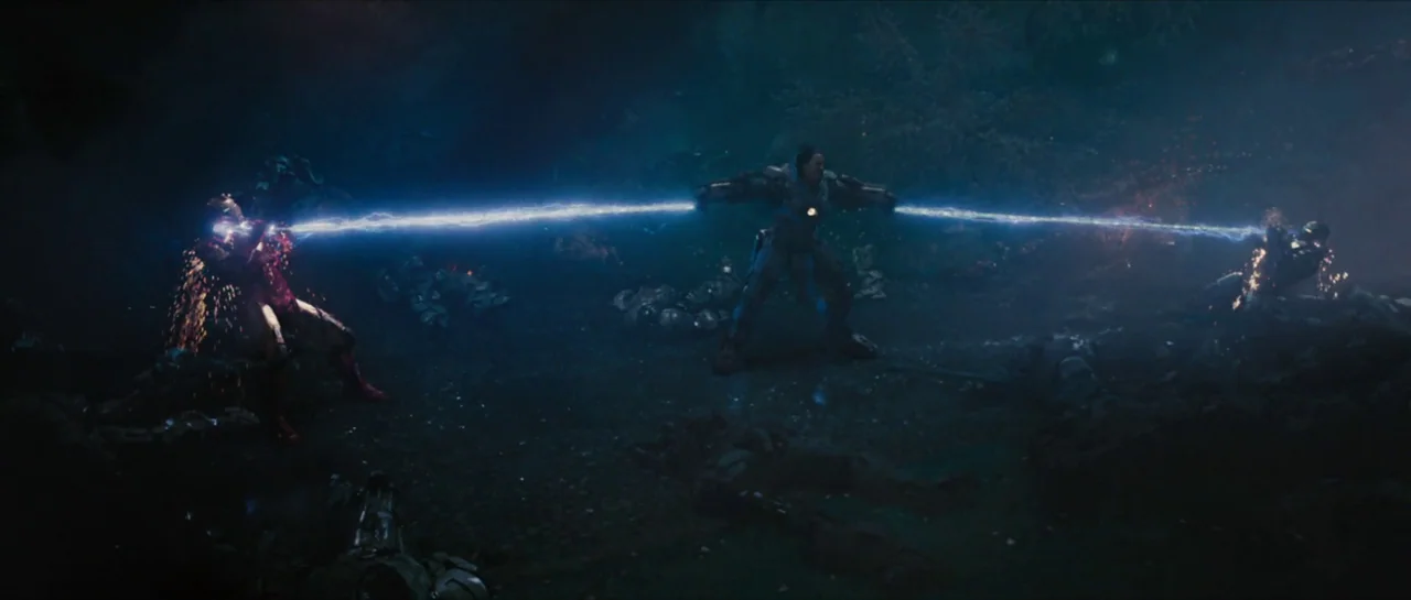 Iron Man 2 (2010) movie still: wide — A dark, debris-strewn ground littered with fallen combatants lies under a night sky. Three figures in advanced…