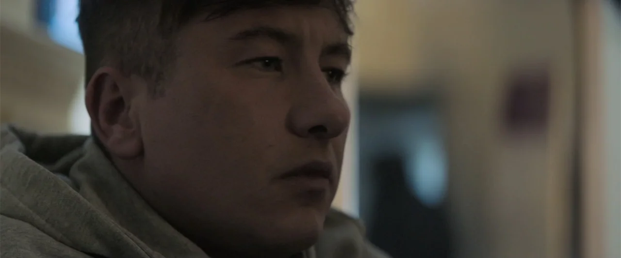 Bring Them Down (2025) movie still: closeup — A man stands in an out-of-focus interior room with blurred architectural shapes behind him. The man is…