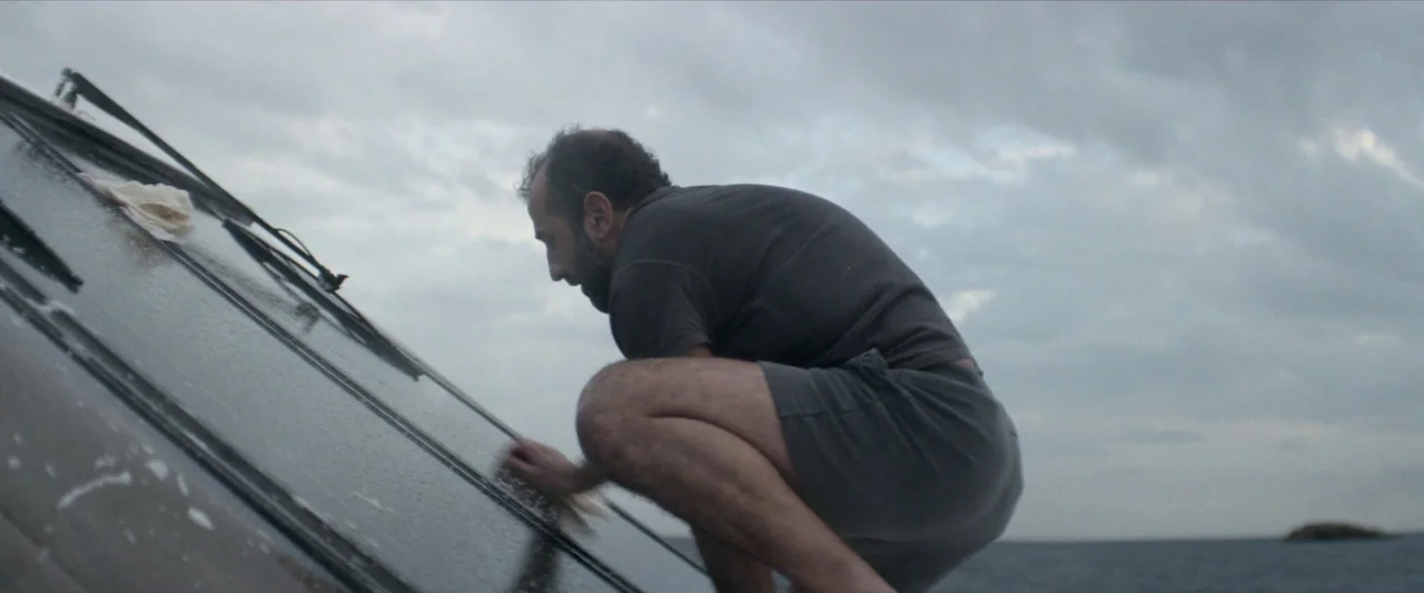Chevalier (2015) movie still: medium — A man crouches on the slanted deck of a boat under a cloudy sky. He cleans a large glass window surface with…