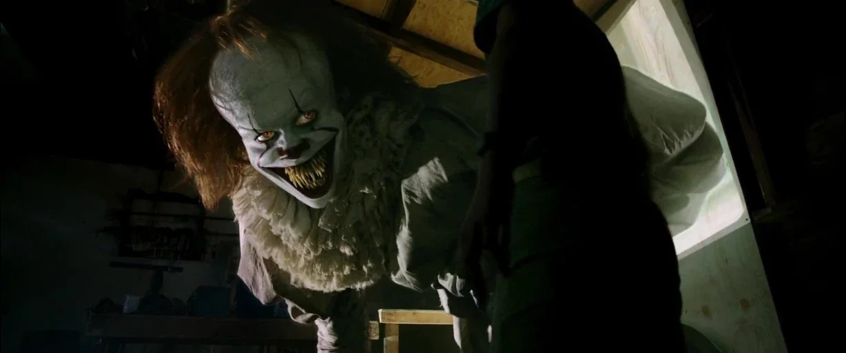 It (2017) movie still: medium — A dark garage interior with wooden beams and cluttered tools visible in the background. A supernatural clown…