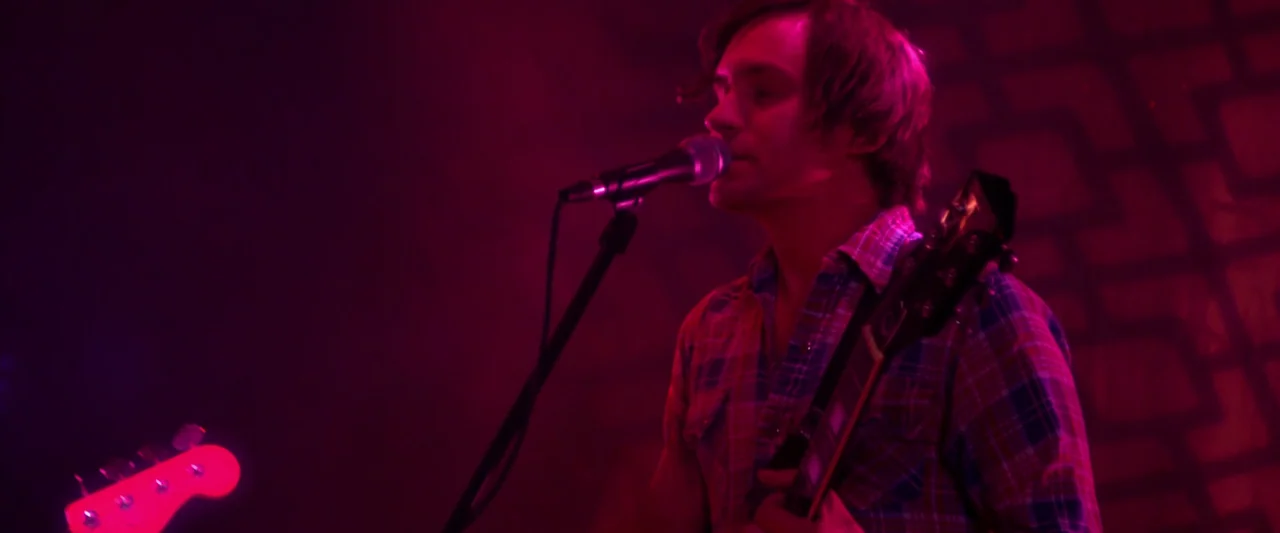 Kaboom (2010) movie still: medium — A man performs on a stage bathed in intense magenta-colored concert lighting. He sings into a microphone while…