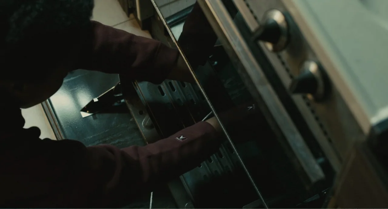 Lila & Eve (2015) movie still: medium — A kitchen interior shows a person wearing a maroon hoodie facing a stainless steel oven. The individual…