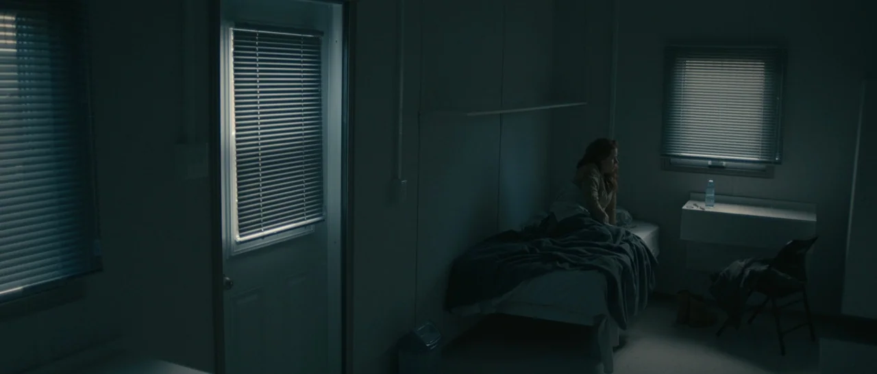 Arrival (2016) movie still: wide — A dimly lit room with plain walls and two windows featuring horizontal blinds. A person sits on the edge of a bed…