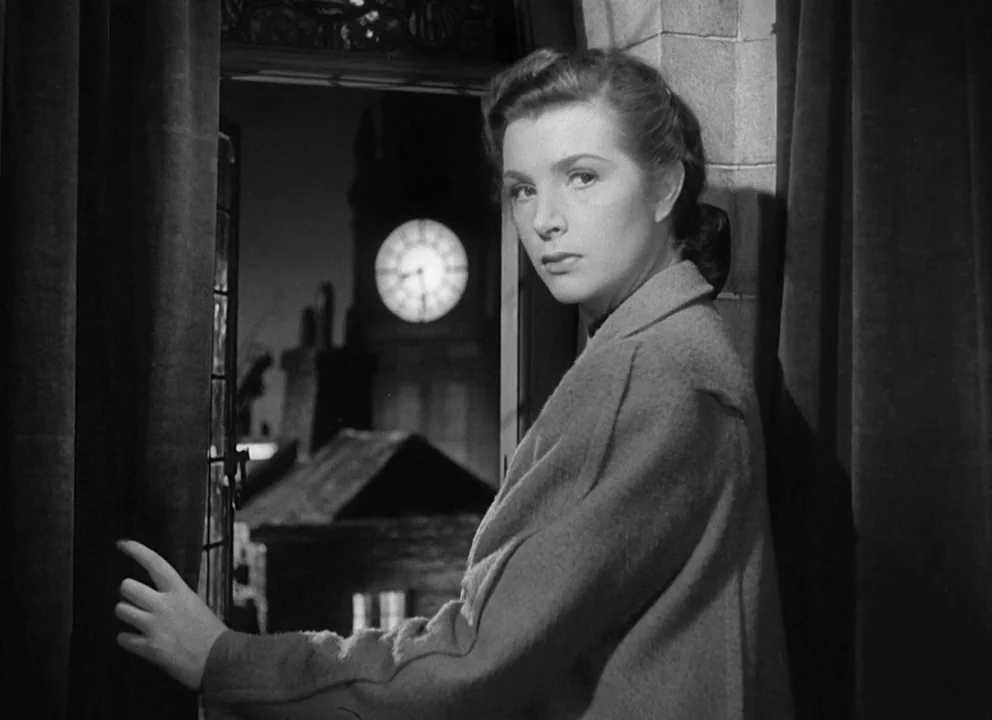 Odd Man Out (1947) movie still: medium — A stone-walled interior next to a window overlooking a night cityscape with a clock. A young woman in a…