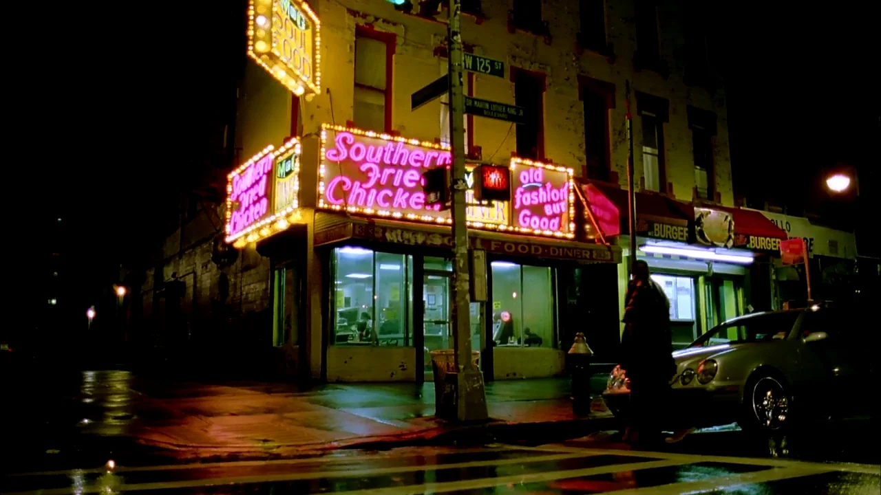 Belly (1998) movie still: wide — A corner diner at night features glowing pink and yellow neon signage on its facade. A silver car is parked near…