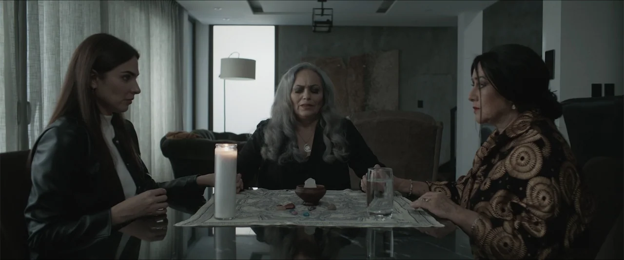 Los habitantes (2023) movie still: medium — A modern interior with neutral walls and soft window light illuminating a dining table. Three women hold…