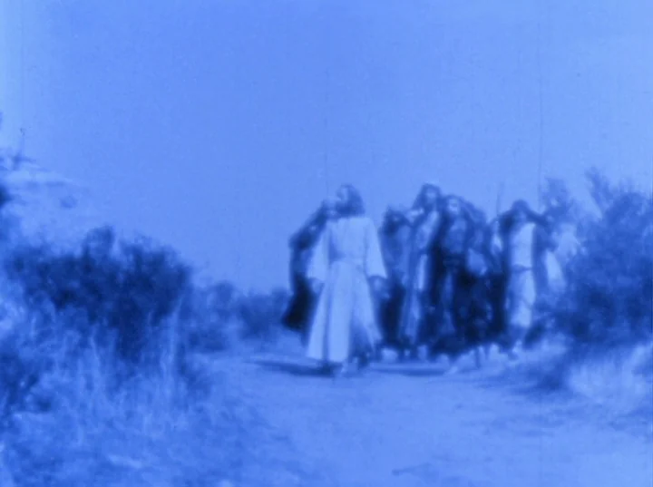 Scorpio Rising (1963) movie still: wide — A group of figures in robes walk along a dusty trail through sparse vegetation. The central figure leads…