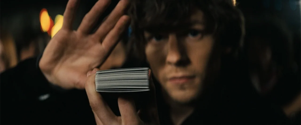 Now You See Me (2013) movie still: closeup — A dark, blurred interior space provides a neutral background for a close-up shot. A young man holds a…