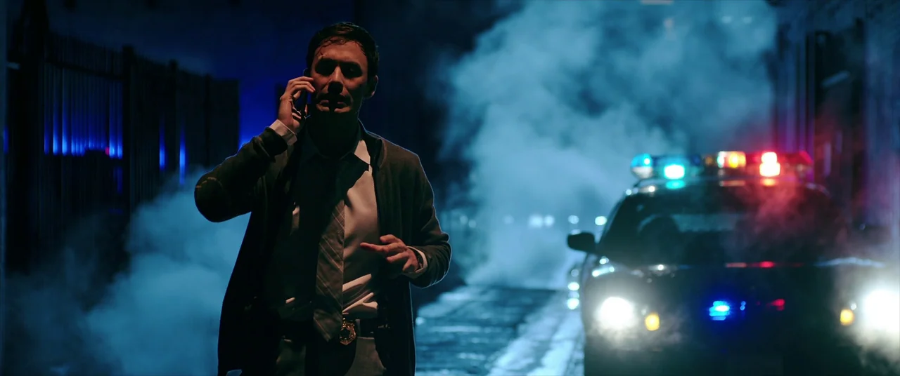 Malignant (2021) movie still: medium — A dark urban alley is filled with thick ground fog and blue ambient light. A man wearing a badge stands…