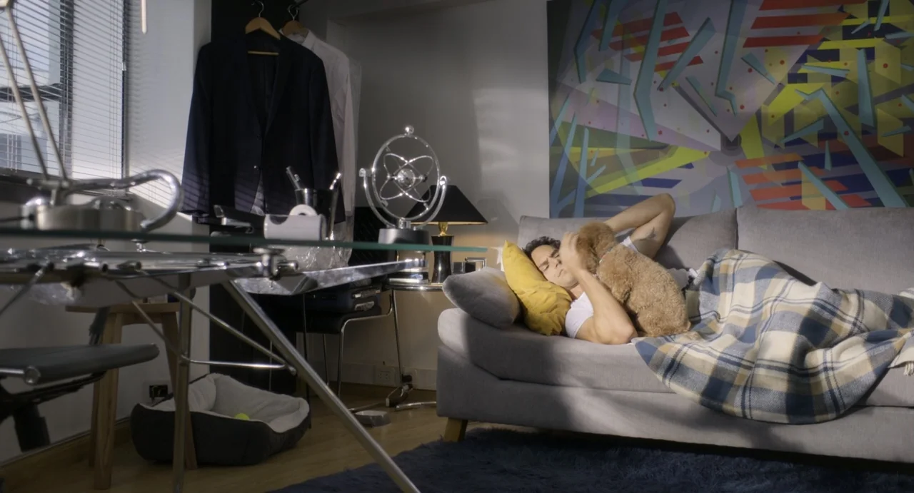 Little White Lies (2010) movie still: medium — A modern living room features a grey couch, abstract wall art, and glass furniture. A young man rests…