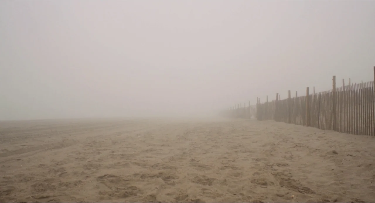 Like Me movie still: establishing — A sandy beach is obscured by a thick layer of dense grey fog. A wooden picket fence runs along the right side…