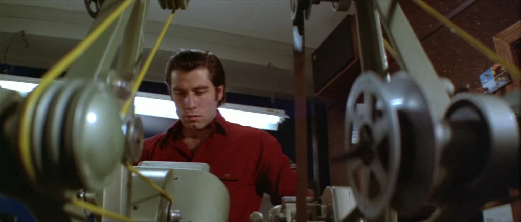 Blow Out (1981) movie still: medium — A man works in a small, cluttered film lab filled with mechanical editing equipment. He sits behind a complex…