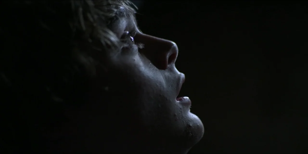 Bring Her Back (2025) movie still: closeup — A dark, undifferentiated interior space obscures the background. A young man looks upward with his face…