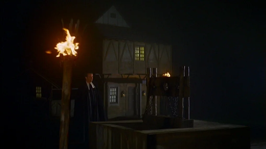 The Scarlet Letter (1973) movie still: wide — A dark, dimly lit village square features a large wooden stocks in the foreground. A man in a dark…