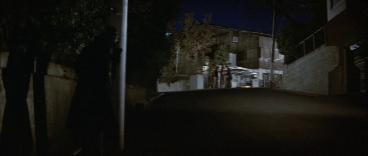 Godzilla Vs Mechagodzilla (1974) movie still: wide — A dark residential street at night featuring a sloping paved road and a wooden house. A…