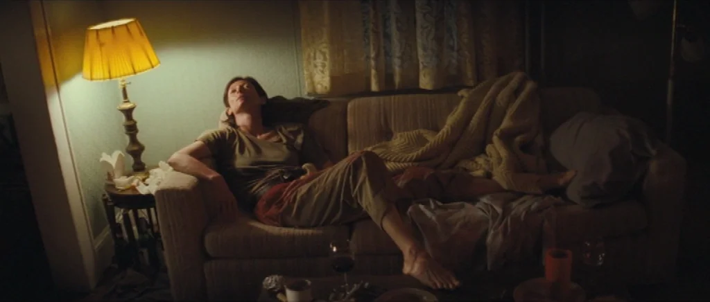 We Need To Talk About Kevin (2011) movie still: medium — A dim living room features a person reclining on a sofa under a lamp. The room contains a…