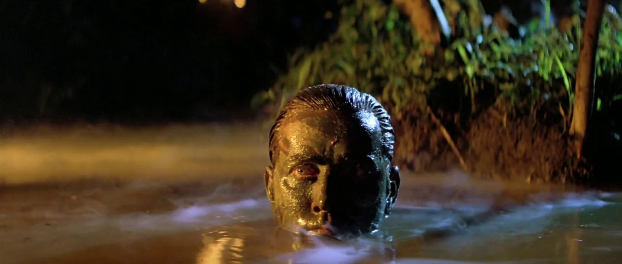 Apocalypse Now (1979) movie still: closeup — A dark river bank at night features lush vegetation and murky water surface. A man's face covered in…