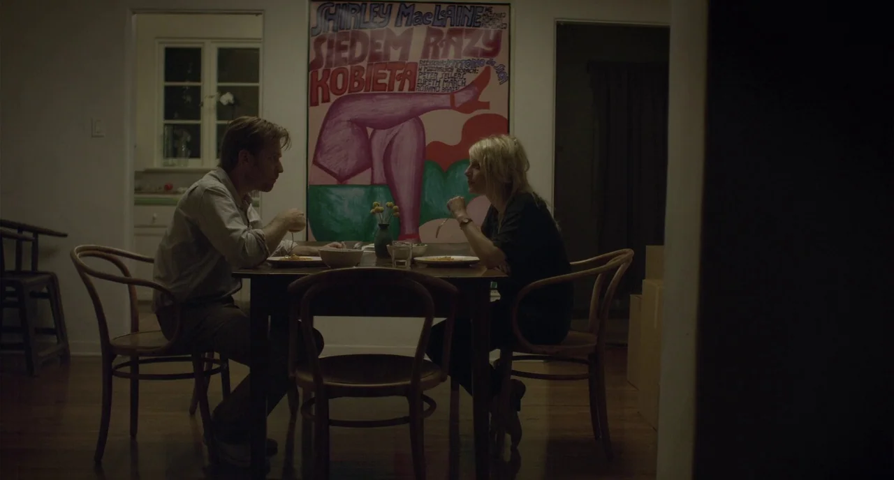 Beginners (2010) movie still: medium — A dimly lit room features a wooden dining table and a large vintage movie poster. Two people sit across from…