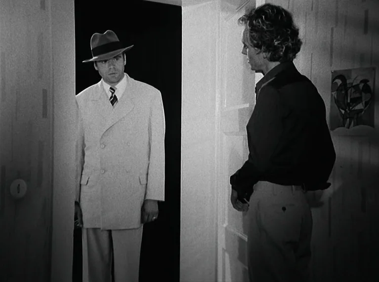 The American Soldier (1970) movie still: medium — A man stands in a dark doorway frame wearing a hat and a light suit. A second man with curly hair…
