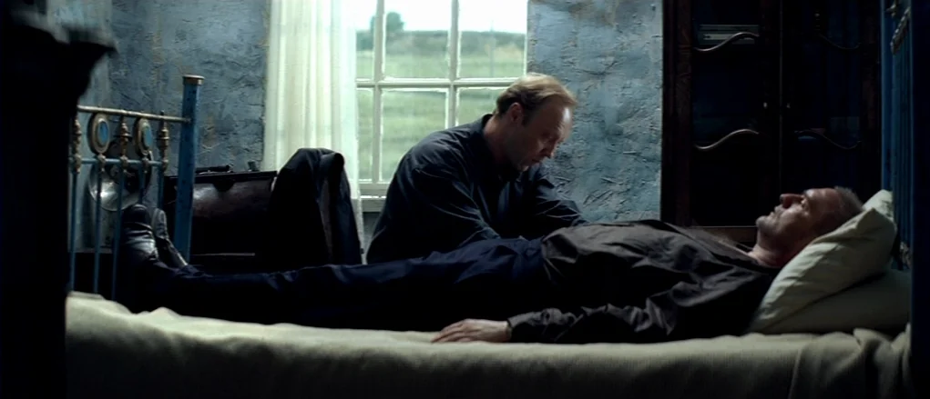 The Banishment (2007) movie still: medium — A dimly lit room with textured blue walls and a window overlooking a field. One man sits by the bedside…