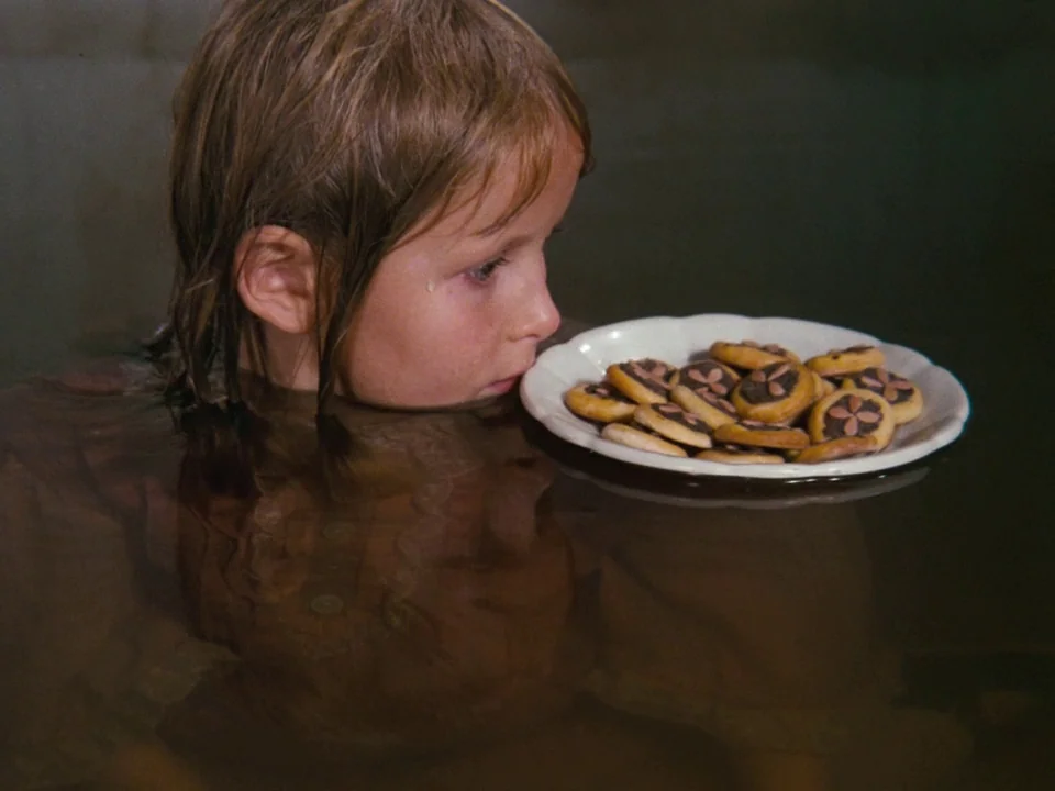 Alice (1988) movie still: closeup — A child rests their chin near a white plate filled with small cookies. The plate floats on the surface of dark,…