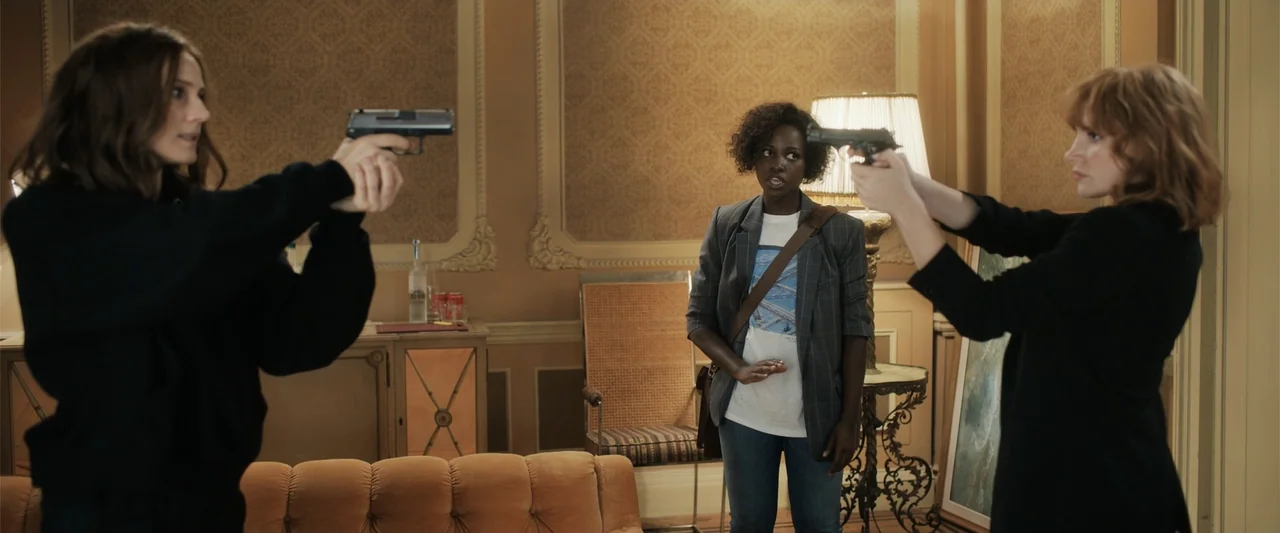 The 355 (2022) movie still: medium — An opulent hotel room with ornate wallpaper and a button-tufted orange couch. Two women aim pistols at each…