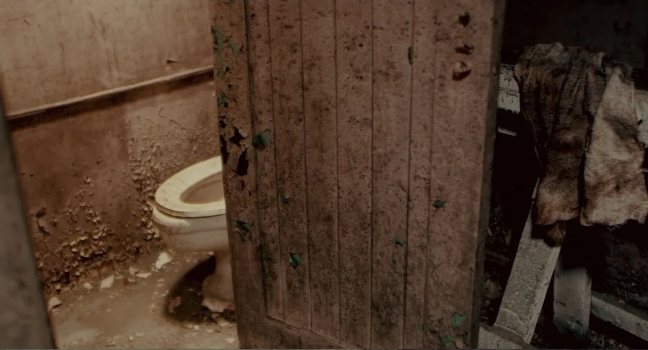Terrifier (2018) movie still: medium — A decrepit restroom stall features walls with peeling paint and a stained concrete floor. A white toilet sits…
