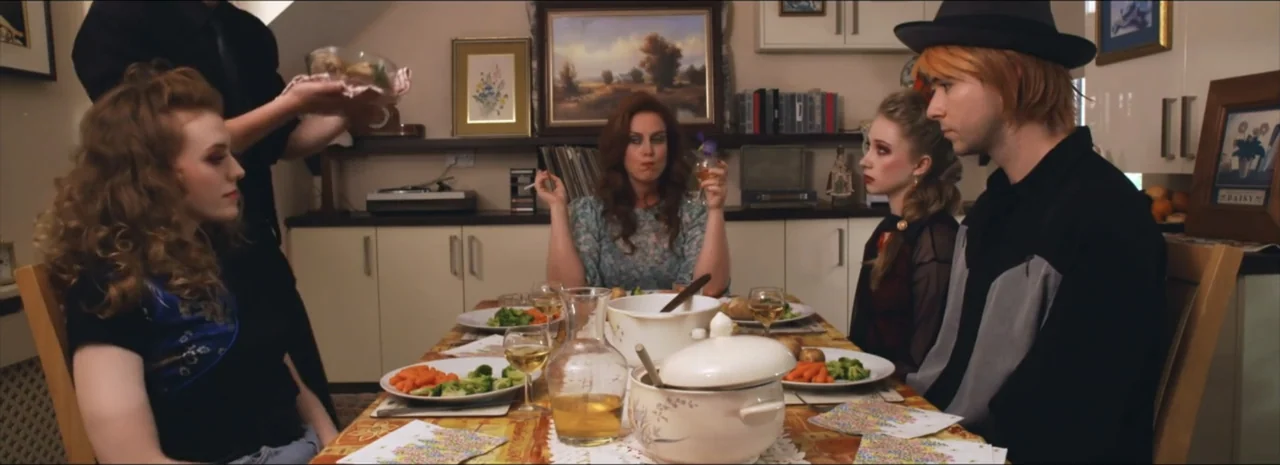 DannyBoy (2020) movie still: medium — A group of people sit at a wooden dining table covered with dishes and food. The individuals are in the middle…