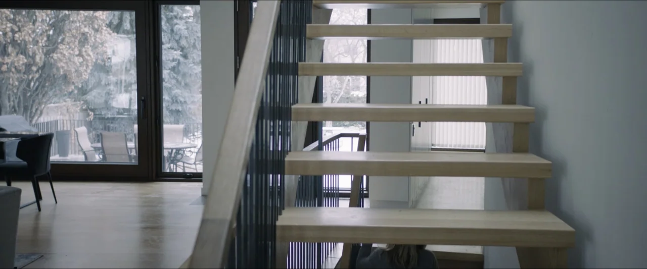 The Lie (2018) movie still: medium — A modern interior staircase with light wood treads and vertical black metal railings. A person stands at the…