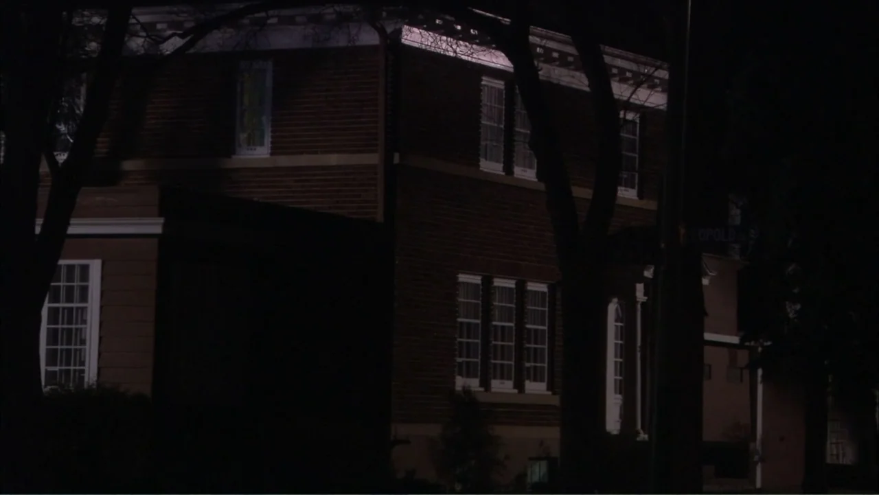 O código oculto (2025) movie still: wide — A multi-story red brick house stands at night behind dark, silhouetted tree trunks. No people are visible…