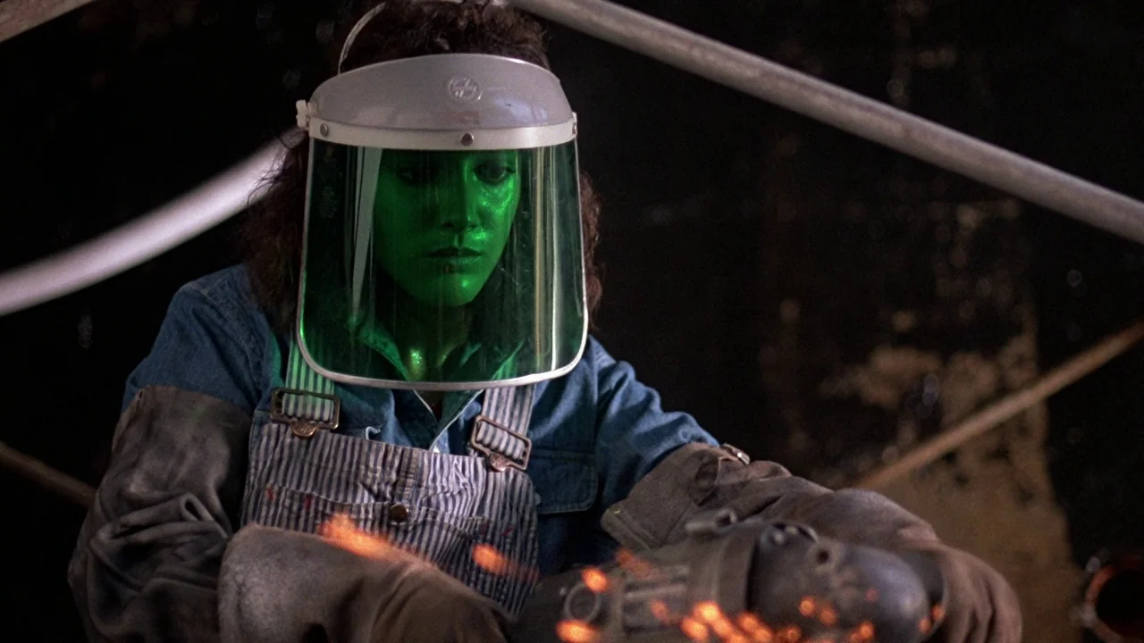 Flashdance (1983) movie still: medium — A dark workshop interior features a person wearing heavy protective gear and a green face shield. The…