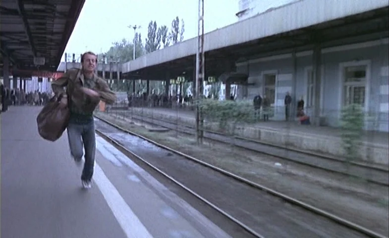 Blind Chance (1987) movie still: fullbody — An outdoor train station platform with concrete flooring and parallel railway tracks. A man runs towards…