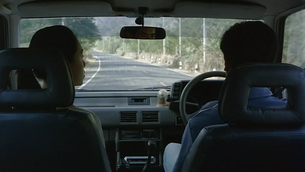 Syndromes and a Century (2006) movie still: medium — The interior of a moving car seen from the back seat, showing two people. A paved road winds…