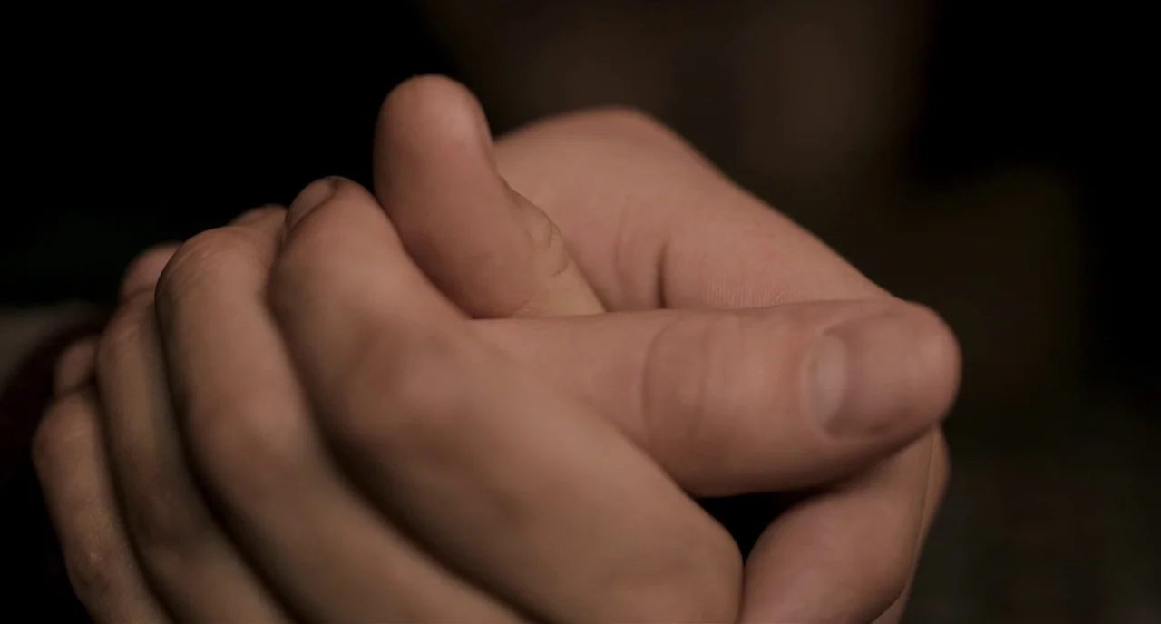 The Selfish Giant (2013) movie still: closeup — Two hands are intertwined with fingers locked together against a dark, featureless background. The…