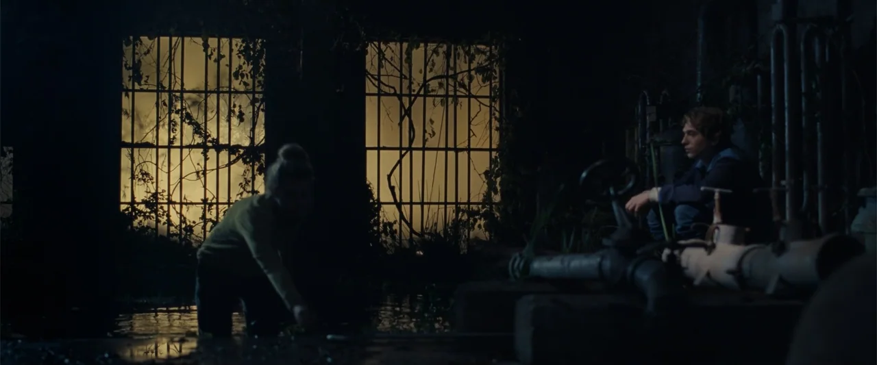 Chemical Hearts (2020) movie still: wide — A dark flooded industrial space contains rusty pipes and two large barred windows. A woman reaches toward…
