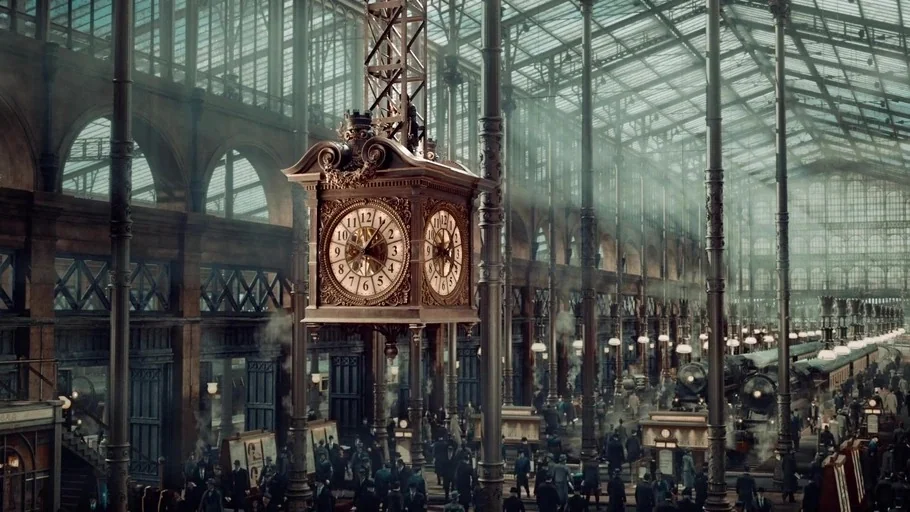 Hugo (2011) movie still: establishing — A grand, high-ceilinged train terminal features ornate iron supports and a sprawling glass roof. A large,…