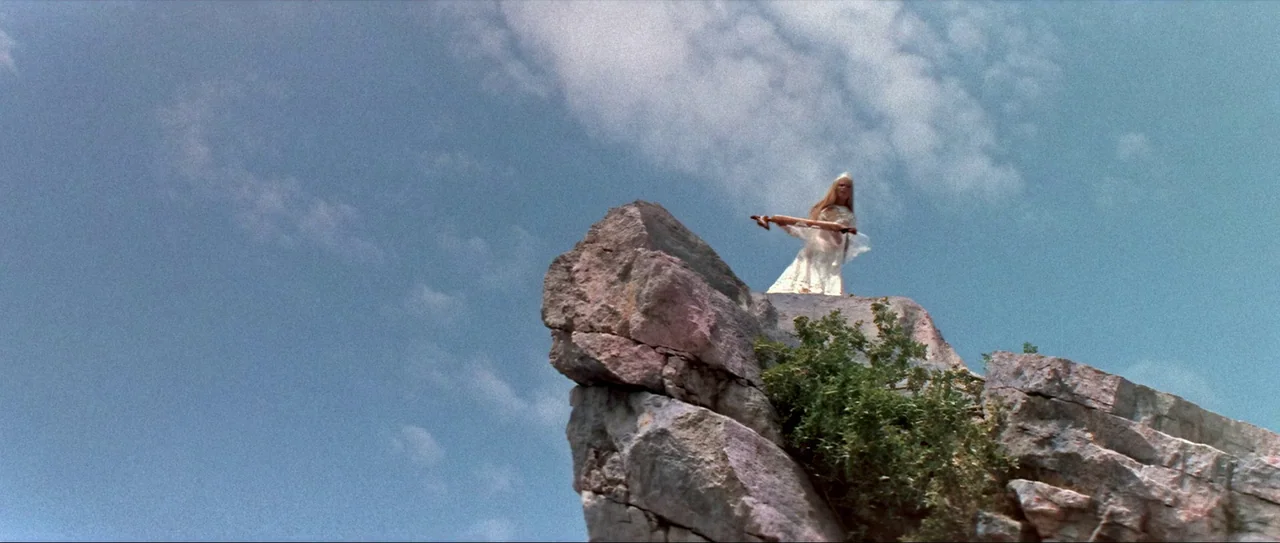 Through Fire, Water and… Brass Pipes (1968) movie still: wide — A high, jagged cliff peak rises against a clear blue sky with thin clouds. A woman…