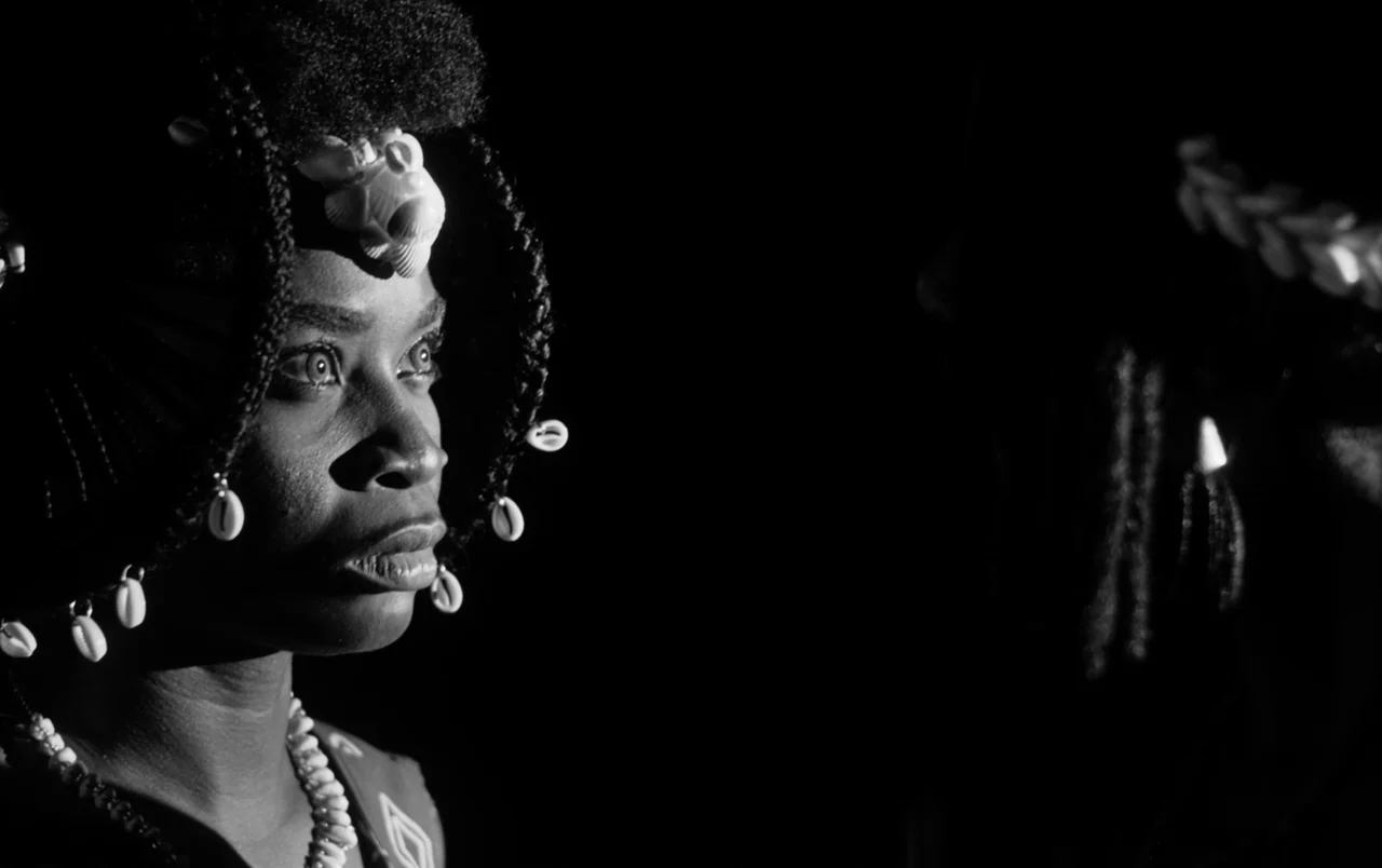 Mami Wata (2023) movie still: closeup — A woman is framed against a void-like black background. She wears braided hair decorated with cowrie shells…