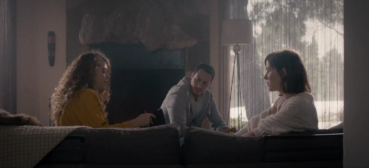 Housewife (2017) movie still: medium — Three individuals sit on couches in a dimly lit room with a fireplace backdrop. They are engaged in a quiet…