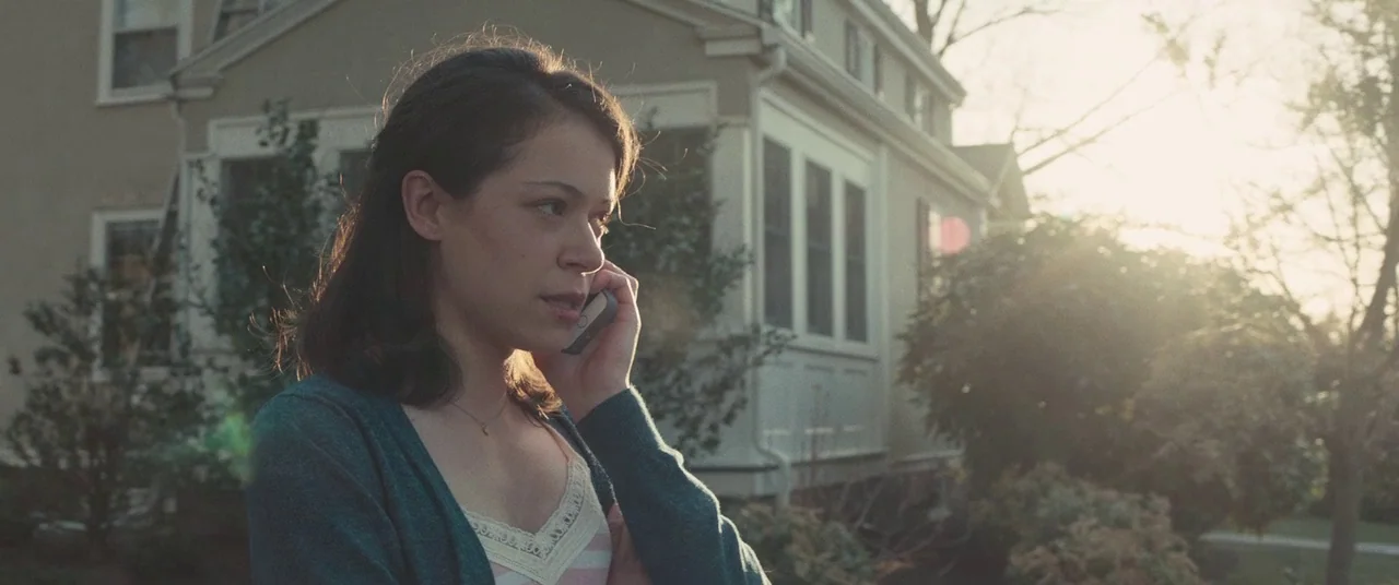 Stronger (2017) movie still: medium — A woman stands in front of a tan two-story house with a white porch. She holds a smartphone to her ear while…