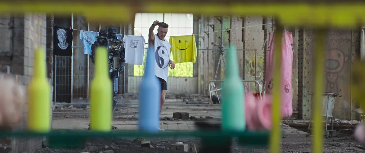 Chappie (2015) movie still: wide — An abandoned industrial warehouse space features exposed steel beams and graffiti-covered brick walls. A man in a…