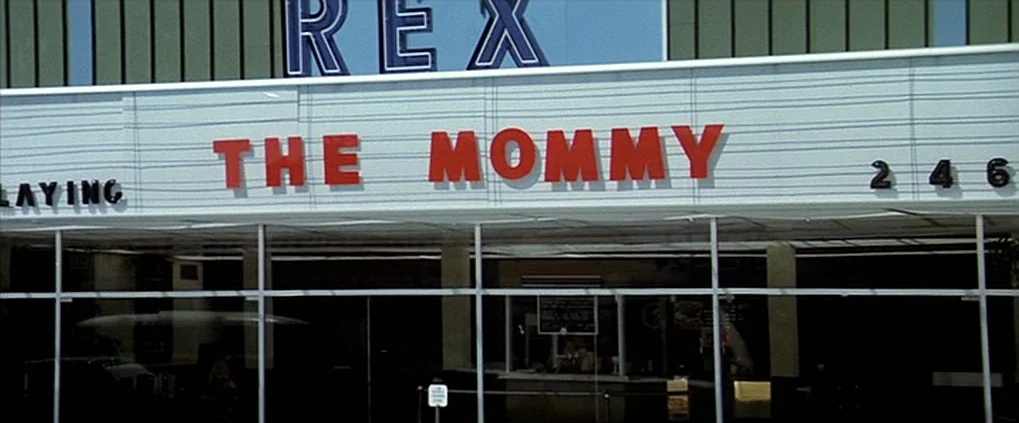 Anguish (1987) movie still: establishing — A 1960s movie theater entrance with a prominent marquee displaying text in red letters. The glass-fronted…