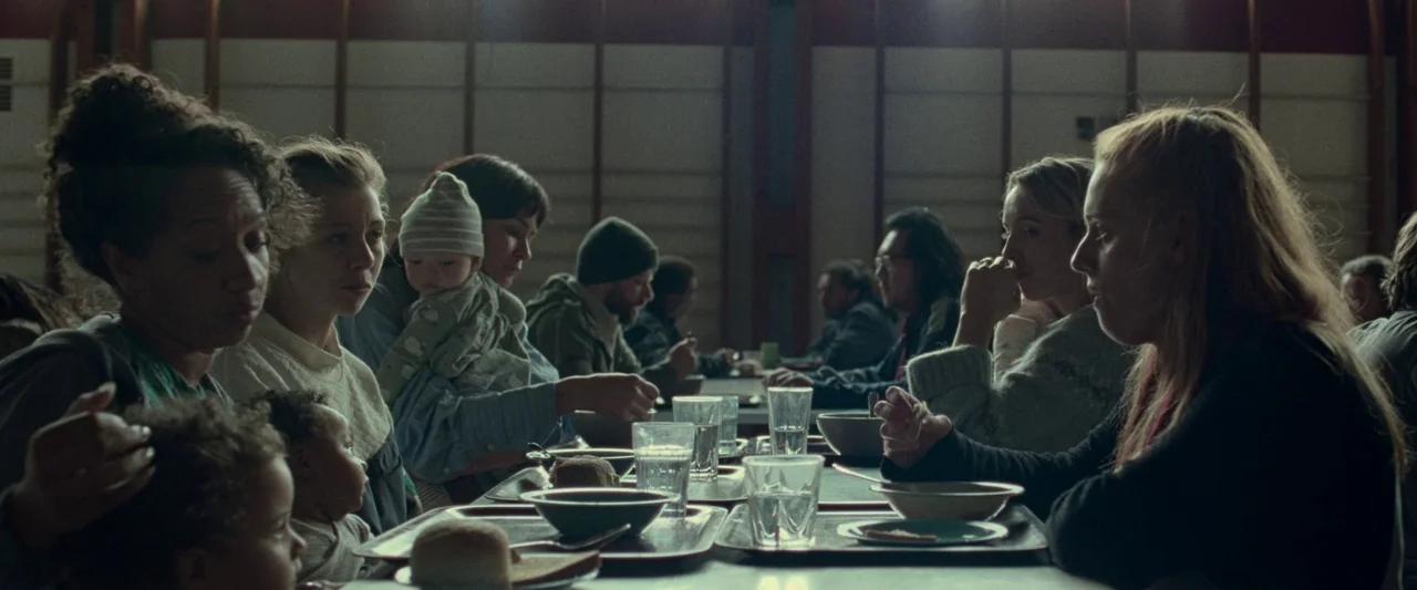 The End We Start From (2023) movie still: wide — A cafeteria interior features long rows of tables with people eating. The lighting is low and…