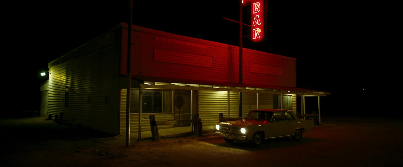 LaRoy, Texas (2023) movie still: wide — A remote roadside building features yellow siding and a prominent red neon bar sign. An older tan sedan is…