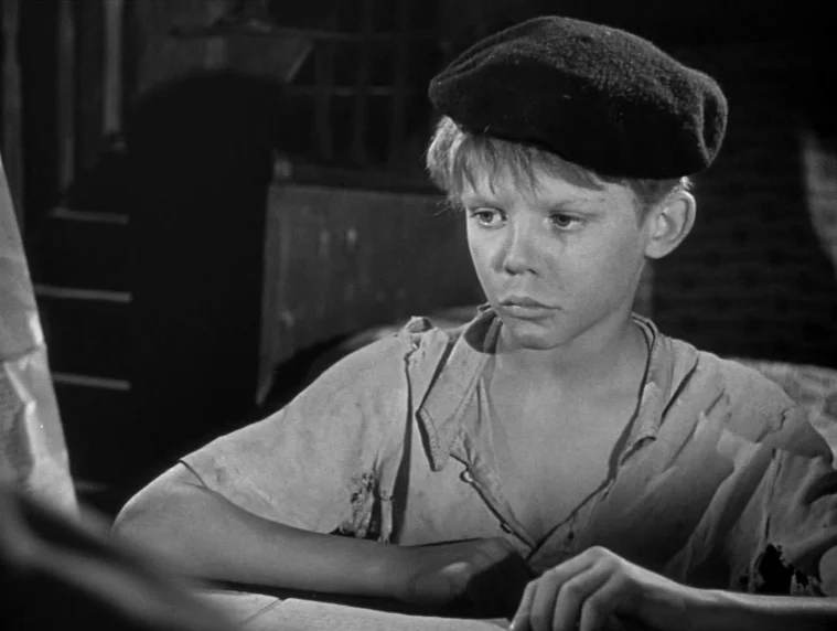 Forbidden Games (1952) movie still: medium — A dimly lit interior room features a young boy sitting at a plain table. He stares towards the side of…