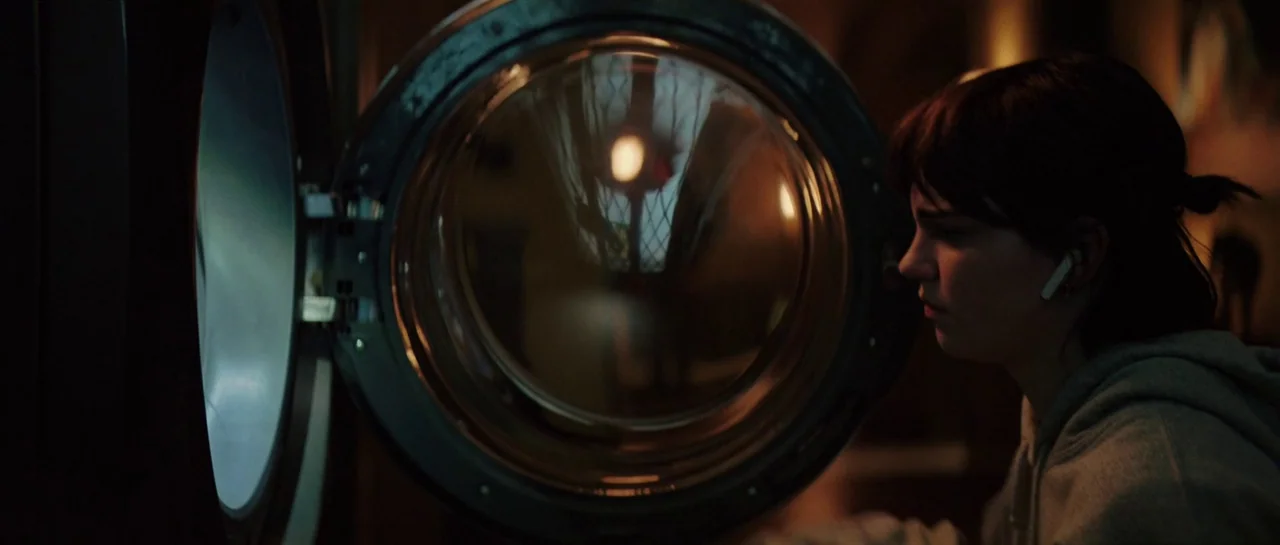 The Boogeyman (2023) movie still: medium — A dark laundry room with a front-loading washing machine in the foreground. A young woman with dark hair…