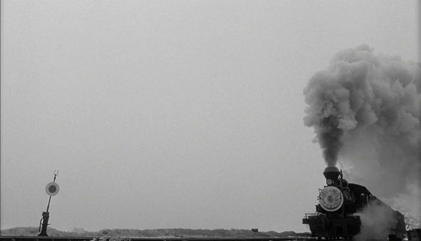 Dead Man (1995) movie still: wide — An old steam locomotive moves across a flat, barren landscape under an overcast sky. A railroad signal stands to…
