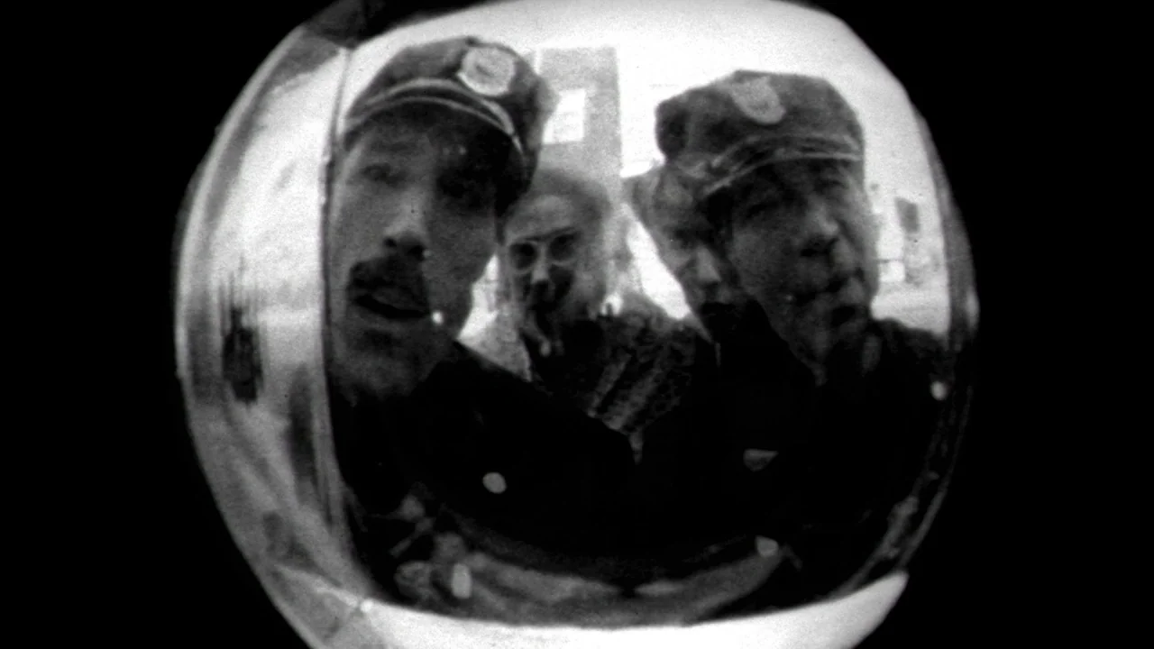 Poison movie still: closeup — A fish-eye view shows several men looking into a door peephole from outside. The men wear peaked caps while peering…